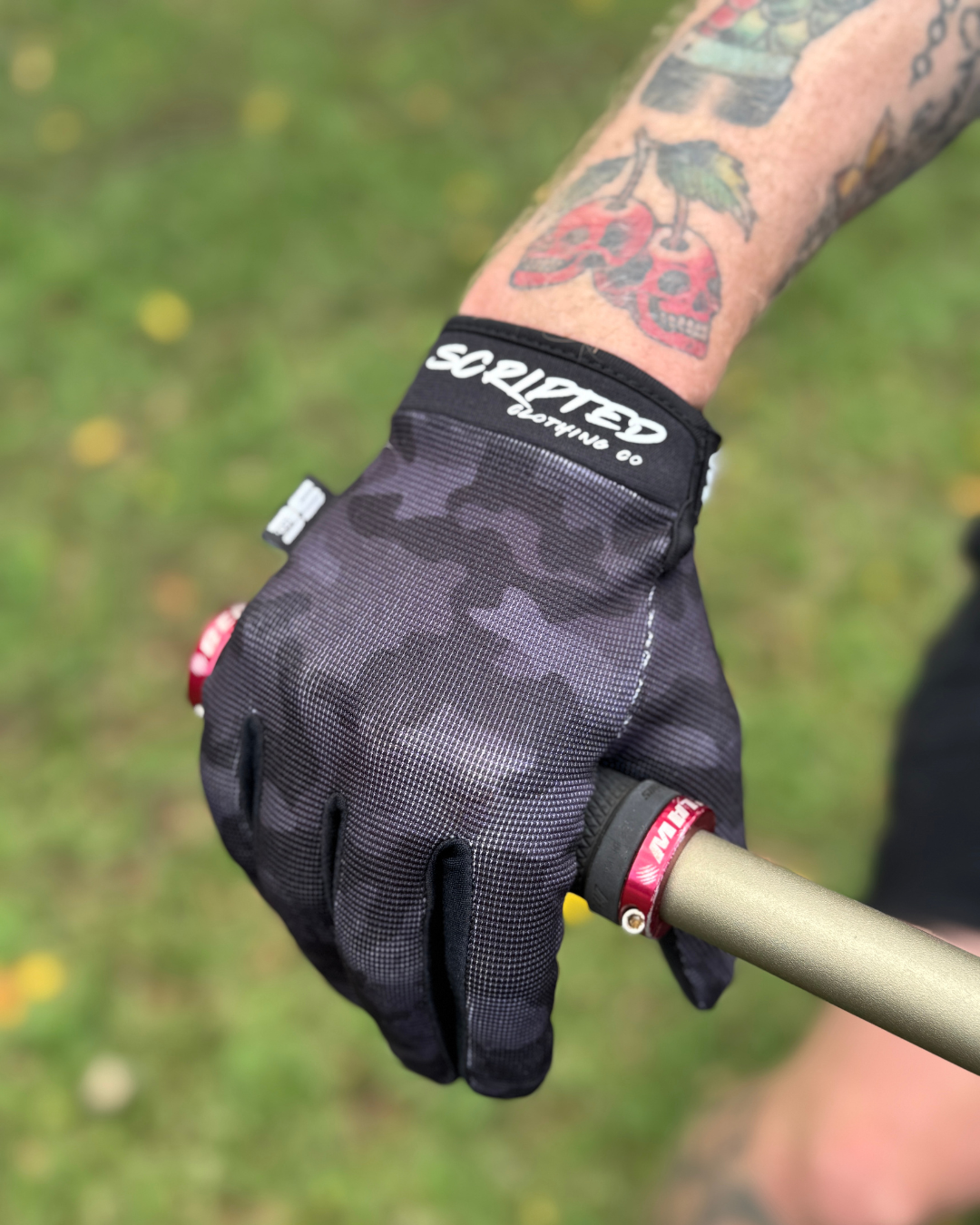 Black camouflage glove on a hand with tattoos, holding onto a BMX bike against a grassy background.